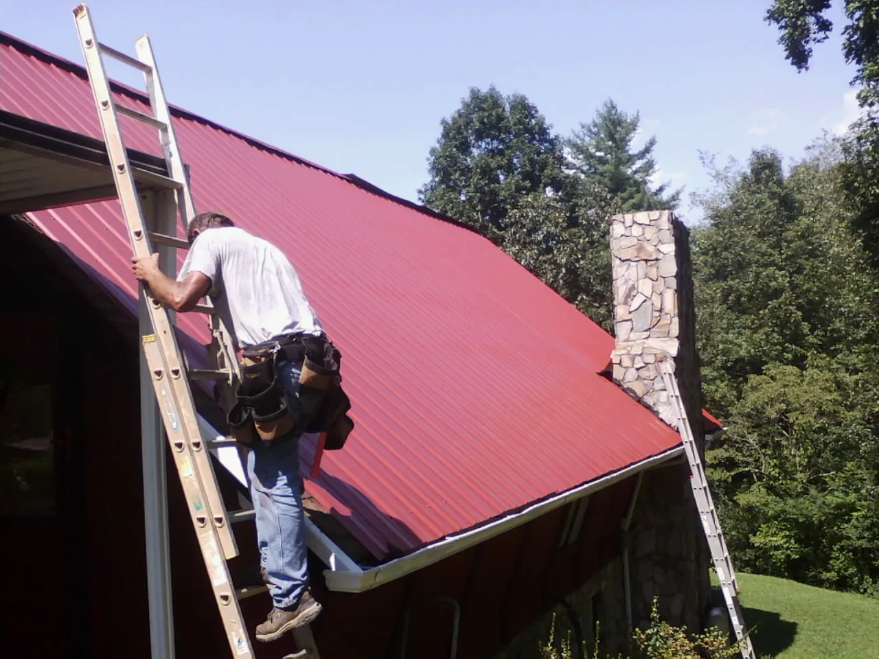 Roofer on a ladder beside a red metal roof and stone chimney — Soffit & Fascia Repair by Roofing Brothers in Norwich